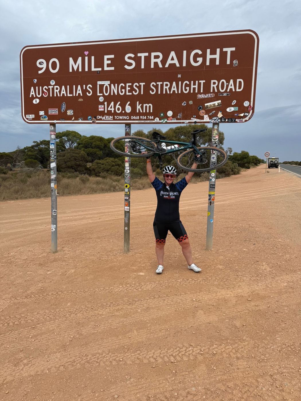 Dawn holding her bike overhead at the 90 Mile Straight sign on the Nullarbor